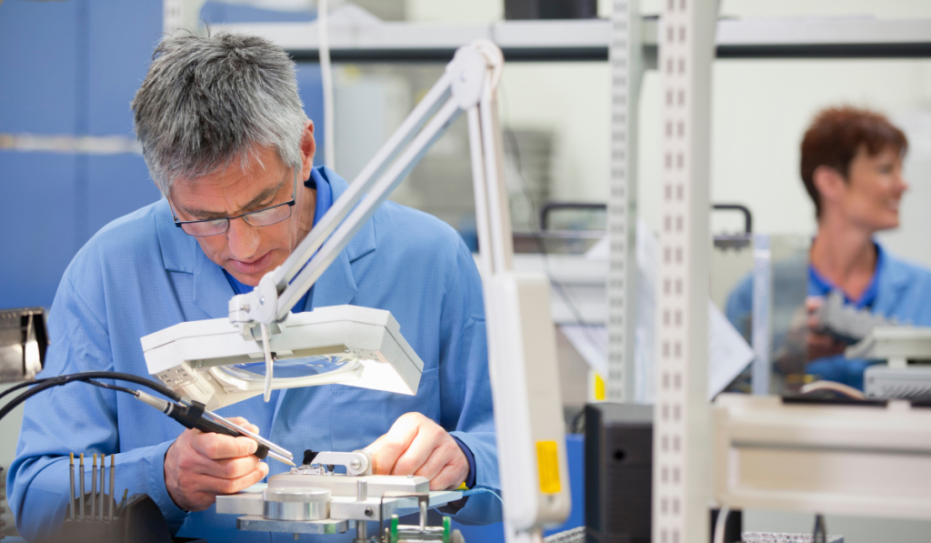 Medical device technician performing precision micro-assembly and inspection under magnification in a controlled manufacturing environment.